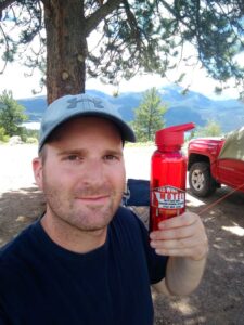 Young man holding Red Wing Motel water bottle at campsite in mountains